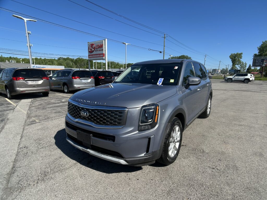 Front three-quarter view of a gray Kia SUV in a dealership lot with other cars under a blue sky.