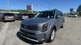 Front three-quarter view of a gray Kia SUV in a dealership lot with other cars under a blue sky.