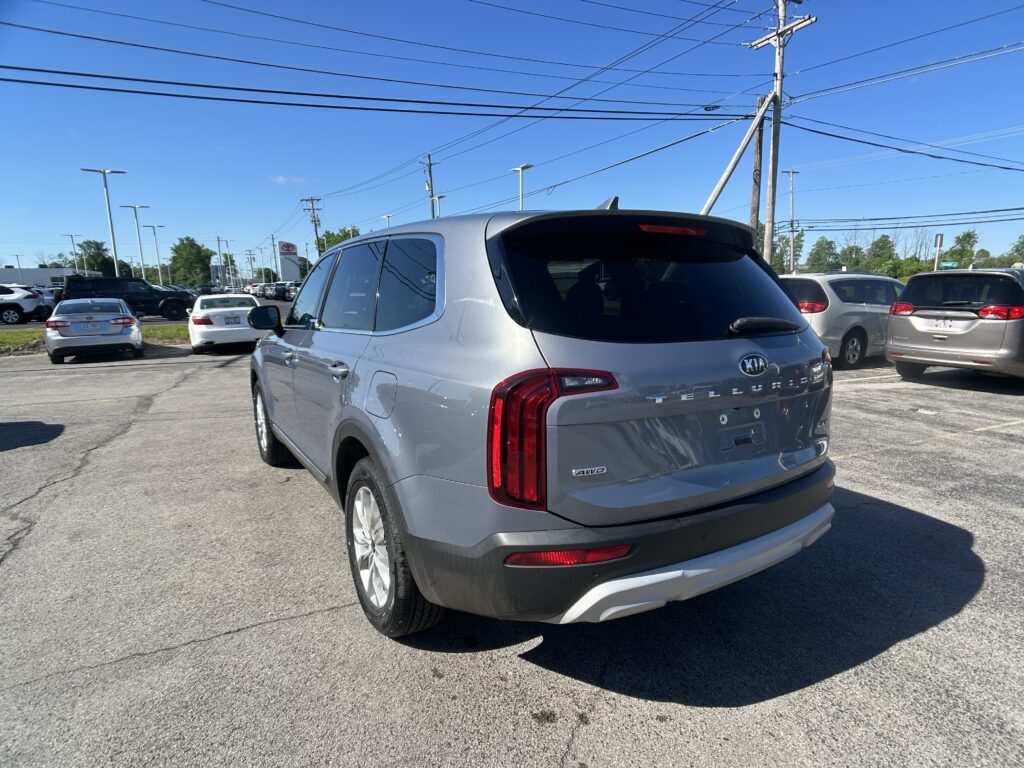 Silver Kia SUV in a busy car dealership lot under a clear blue sky with power lines overhead, rear three-quarter view.
