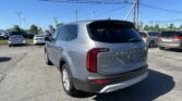 Silver Kia SUV in a busy car dealership lot under a clear blue sky with power lines overhead, rear three-quarter view.
