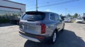 Rear three-quarter view of a gray SUV in a car dealership lot, with a white building and other vehicles in the background, under a bright blue sky.
