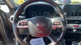 Driver's view of car interior with Kia steering wheel and dashboard, visible dealership parking lot through windshield, outside cars.