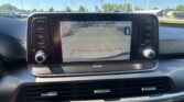 Car infotainment screen showing rear-view camera with guiding lines and the message 'Check surroundings for your safety.' with vehicle controls on left and right and a parking lot outside.