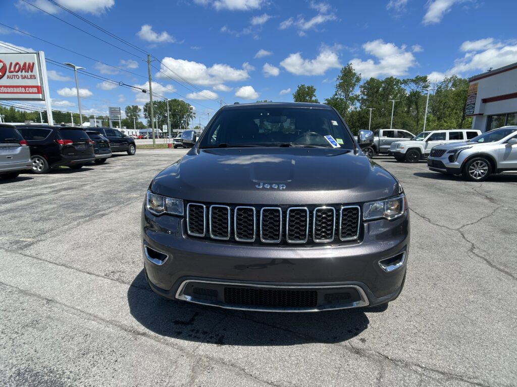 Front view of a dark gray Jeep SUV parked in a dealership lot under a blue sky with other cars around.