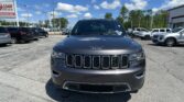 Front view of a dark gray Jeep SUV parked in a dealership lot under a blue sky with other cars around.