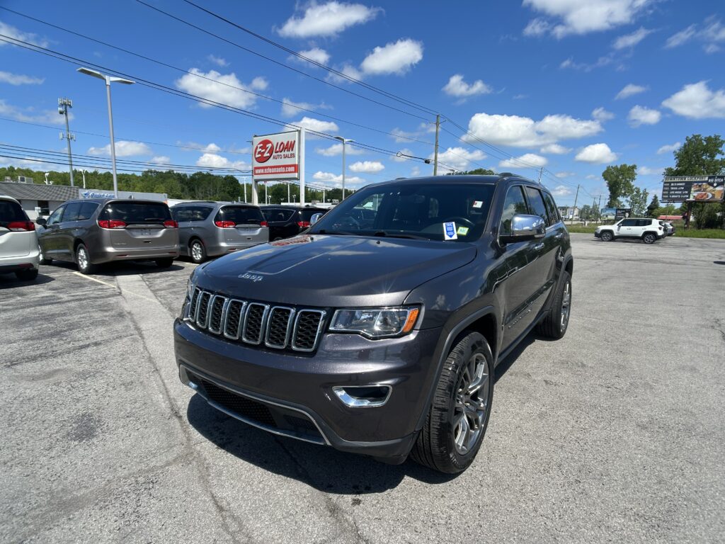 Close-up of a dark gray Jeep SUV in a car dealership lot with other vehicles and a red EZ LOAN sign in the background, blue sky above.