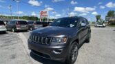 Close-up of a dark gray Jeep SUV in a car dealership lot with other vehicles and a red EZ LOAN sign in the background, blue sky above.