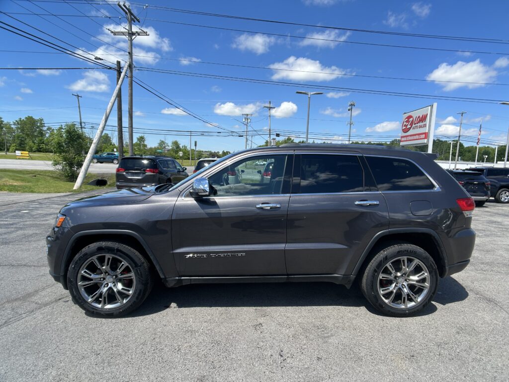 Gray Jeep Grand Cherokee SUV parked in a car dealership lot with an EZ Loan Auto Sales sign in the background and power lines overhead under a blue sky.