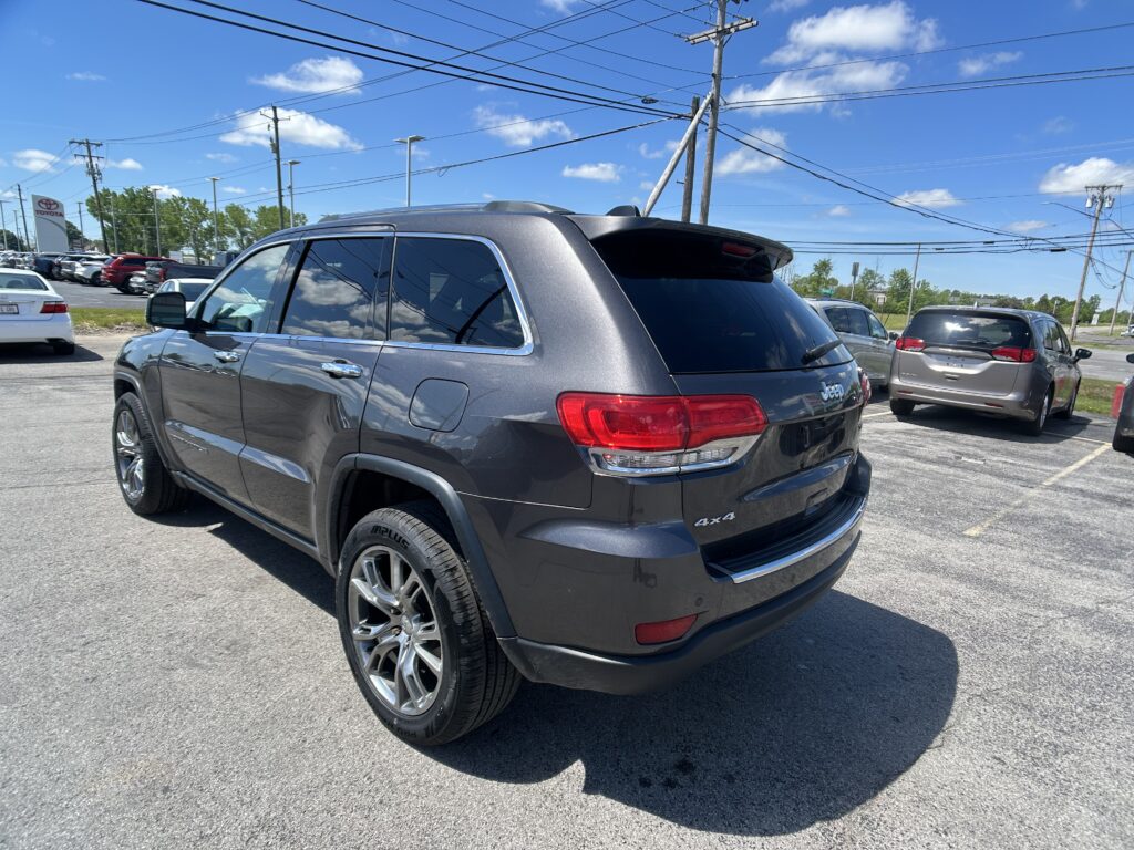 Rear three-quarter view of a gray Jeep Grand Cherokee in a parking lot on a sunny day with power lines overhead and other vehicles nearby.