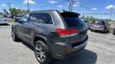 Rear three-quarter view of a gray Jeep Grand Cherokee in a parking lot on a sunny day with power lines overhead and other vehicles nearby.