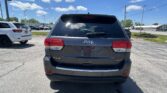 Rear of a gray Jeep SUV (Limited 4x4) in a car dealership lot with other vehicles and power lines, blue sky and clouds above.