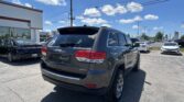 Rear of a dark gray Jeep Grand Cherokee Limited in a car dealership lot, with other cars, Toyota signs in the distance, under a blue sky with clouds and power lines.