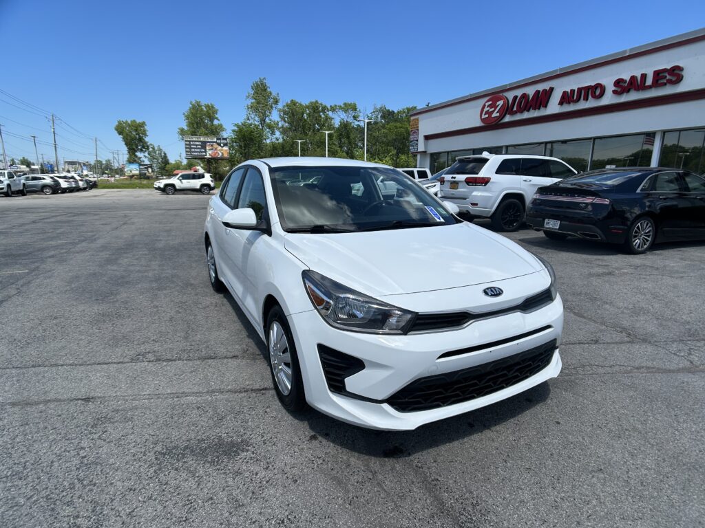 White Kia sedan in the foreground at an auto dealership lot with other cars and a red EZ Loan Auto Sales sign in the background.