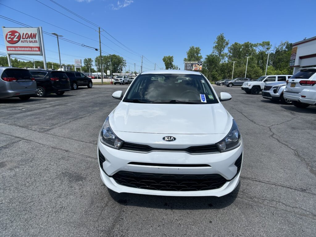 Front view of a white Kia in a parking lot with other cars and a blue sky above, dealership scene in the background.