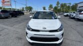 Front view of a white Kia in a parking lot with other cars and a blue sky above, dealership scene in the background.