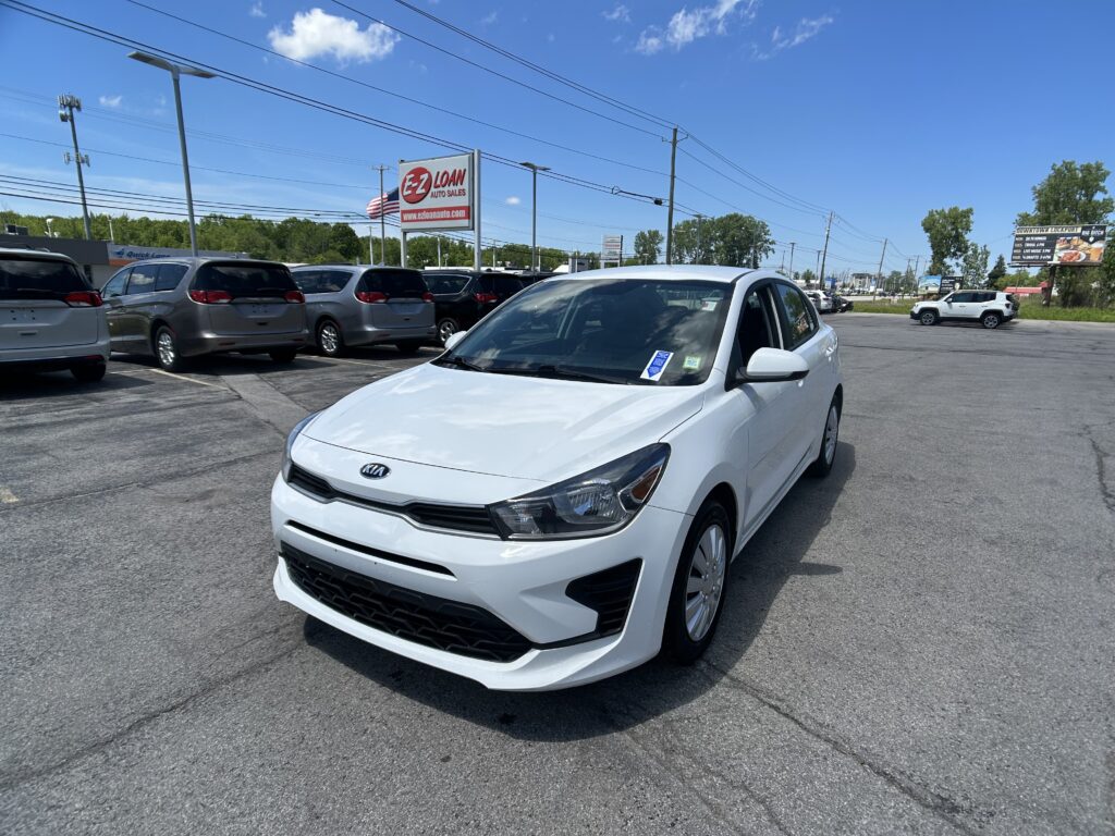 White Kia sedan in the foreground at a car dealership lot, with a row of minivans and an EZ Loan Auto Sales sign against a blue sky.