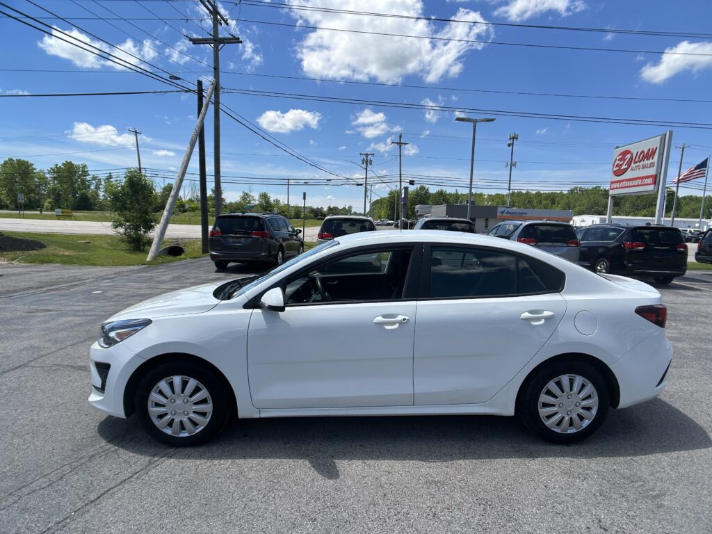 White sedan in a car dealership lot with other vehicles and a tall power pole in the background under a blue sky with clouds