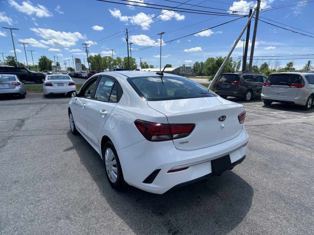 Rear three-quarter view of a white Kia Rio in a car dealership parking lot on a sunny day, with several other cars around and power lines overhead.
