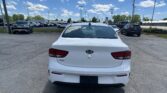 Rear view of a white Kia Rio in a busy dealership parking lot under a bright blue sky with scattered clouds.
