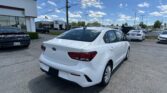 White Kia sedan in the foreground at a car dealership lot with a Toyota dealership in the background and a blue sky above.