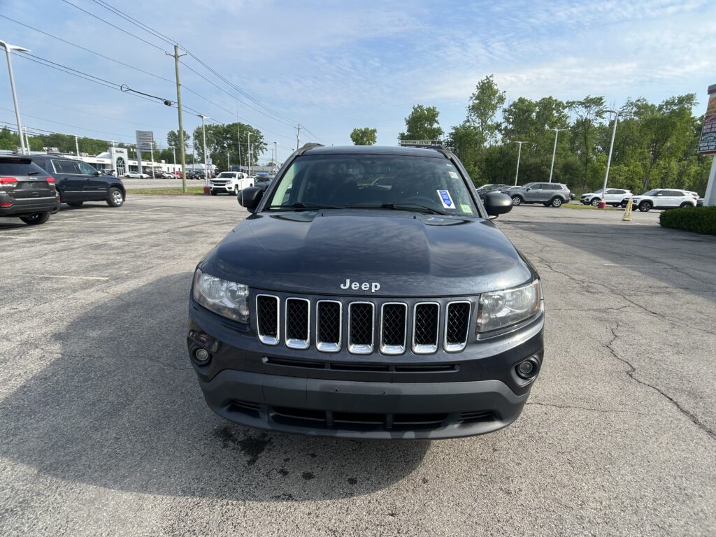Front view of a dark gray Jeep vehicle in a parking lot at a car dealership, with other cars and dealership buildings in the background