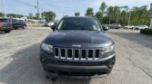 Front view of a dark gray Jeep vehicle in a parking lot at a car dealership, with other cars and dealership buildings in the background