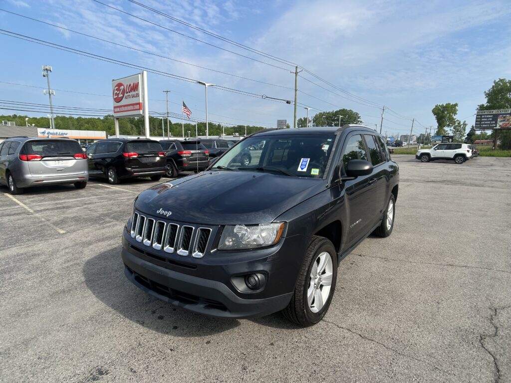 Front-right view of a dark blue Jeep SUV in a car dealership lot, with other vehicles, a tall EZ LOAN Auto Sales sign, and an American flag in the background.
