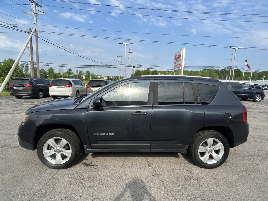 Front-right view of a dark gray Jeep Compass SUV in a dealership lot with other minivans, power lines overhead and a blue sky.