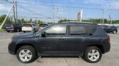 Front-right view of a dark gray Jeep Compass SUV in a dealership lot with other minivans, power lines overhead and a blue sky.