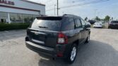 Rear view of a black Jeep SUV in a car dealership lot with other cars and a 'SALES' sign on the building visible in the background