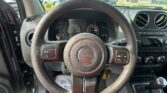 Driver's view of a Jeep steering wheel with dashboard gauges visible and center console electronics in sight, outside cars and buildings through the windshield.