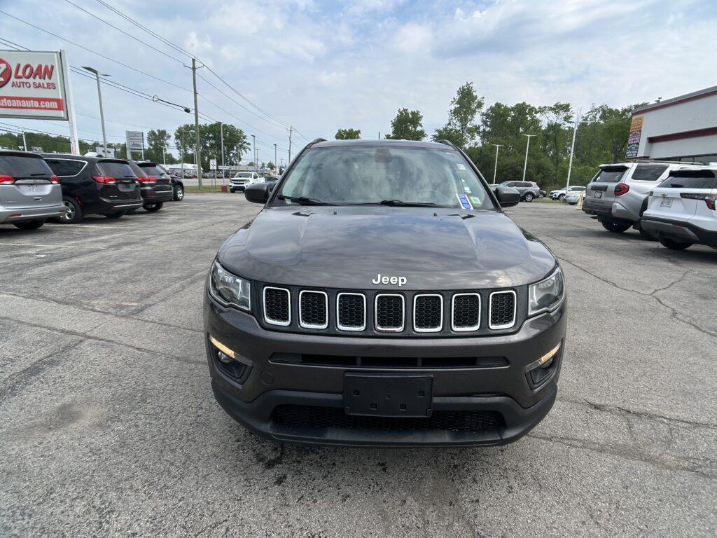 Front view of a gray Jeep SUV in a car dealership lot with other vehicles and signs in the background