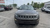 Front view of a gray Jeep SUV in a car dealership lot with other vehicles and signs in the background