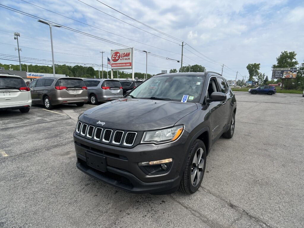 Close-up view of a dark gray Jeep SUV in a dealership lot with a row of minivans behind it and an EZ Loan Auto Sales sign in the distance, under a partly cloudy sky.