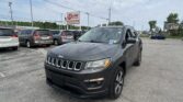 Close-up view of a dark gray Jeep SUV in a dealership lot with a row of minivans behind it and an EZ Loan Auto Sales sign in the distance, under a partly cloudy sky.