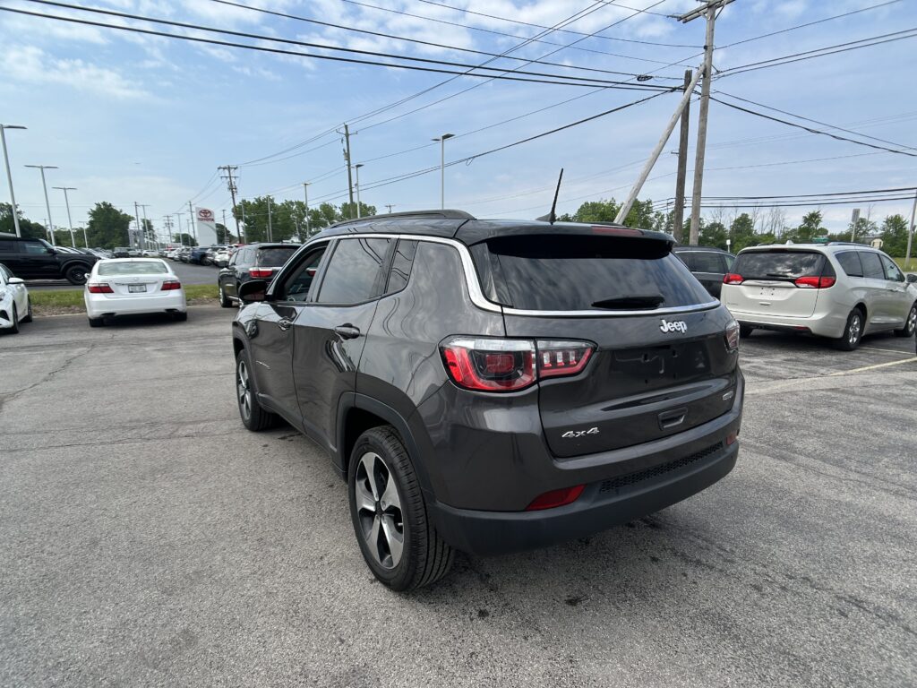 Rear view of a dark gray Jeep SUV in a car dealership parking lot with other cars and crisscrossing power lines overhead against a blue sky.