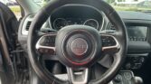 Driver's-seat view of a Jeep interior with steering wheel centered, instrument cluster, and infotainment screen; outdoors visible through windshield in a parking lot.
