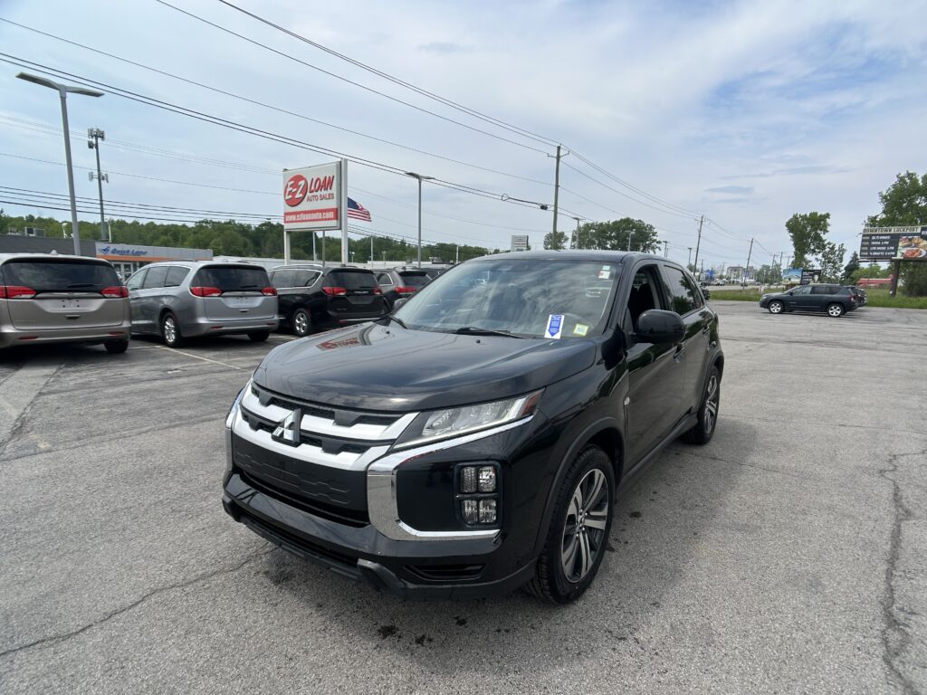 Black SUV in the foreground at a car dealership lot with several vans and a sign reading EZ-Loan Auto Sales in the background, plus an American flag nearby.
