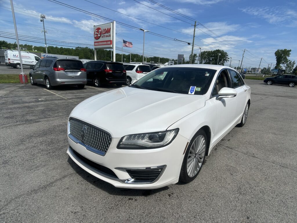 Close-up view of a white sedan in the dealership lot, with other cars and an EZ Loan Auto Sales sign in the background under a blue sky.