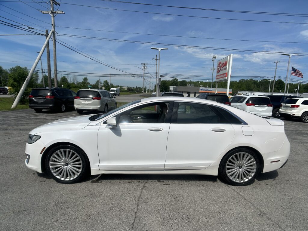 White luxury sedan in foreground at a car dealership lot with large chrome wheels and blue sky above, power lines in the background and an American flag nearby