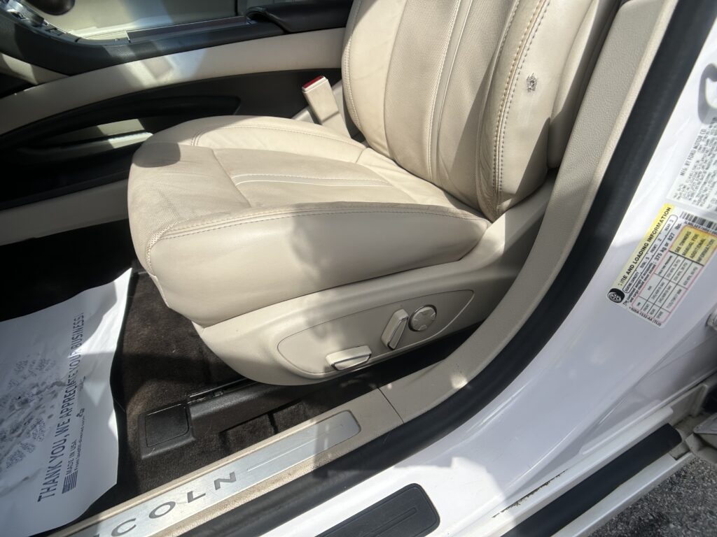 Beige leather car driver seat with power-adjustment controls on the lower side and part of the door frame visible.
