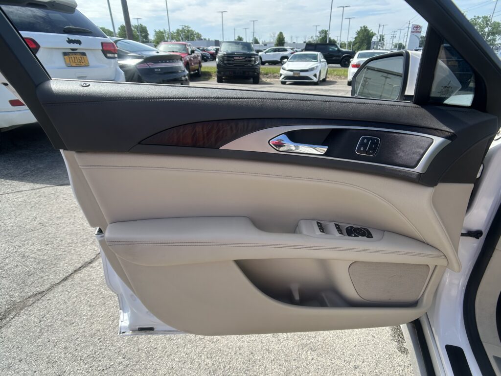 Interior of a white car door: beige leather, wood trim, chrome handle, and window control buttons; view of a busy parking lot outside with several cars.