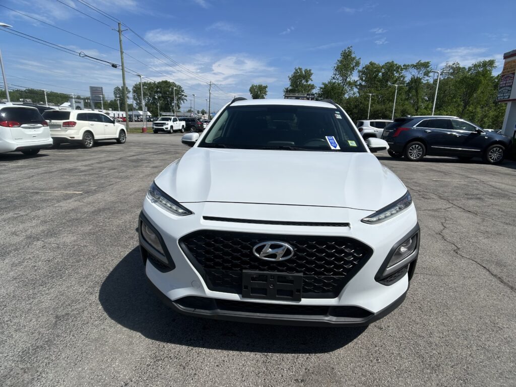 Front view of a white Hyundai SUV in a crowded parking lot on a sunny day, other cars nearby and blue sky above.
