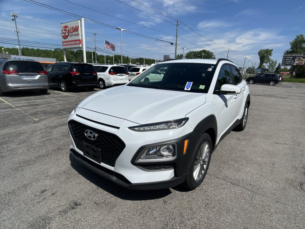 White Hyundai SUV in foreground at a car dealership lot with other vehicles, a sign reading 'EZ LOAN Auto Sales' and an American flag in the background