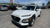 White Hyundai SUV in foreground at a car dealership lot with other vehicles, a sign reading 'EZ LOAN Auto Sales' and an American flag in the background