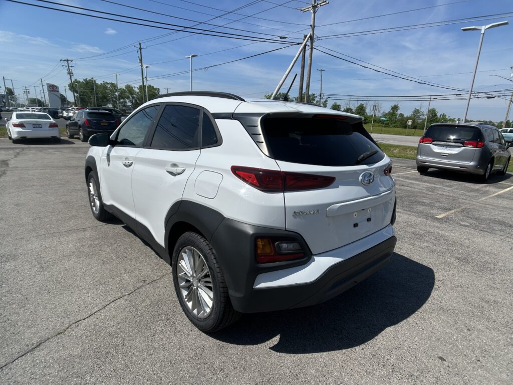 White Hyundai Kona shown in a dealership parking lot, rear three-quarter view with other cars and power lines overhead on a sunny day.