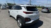 White Hyundai Kona shown in a dealership parking lot, rear three-quarter view with other cars and power lines overhead on a sunny day.