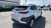 White Hyundai SUV parked in a car dealership lot, storefront showing a large 'SALES' sign to the left and other vehicles in the background to the right.