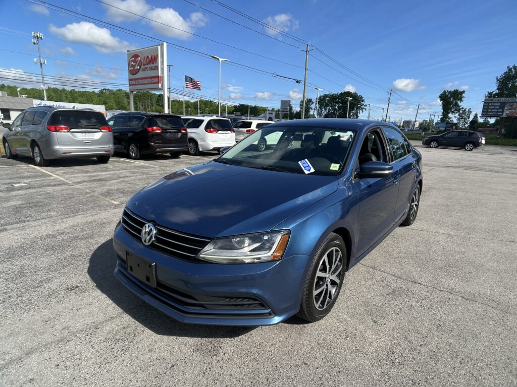 Blue Volkswagen sedan in the foreground at a car dealership lot with several minivans and an EZ Loans Auto Sales sign nearby, American flag in background.