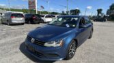 Blue Volkswagen sedan in the foreground at a car dealership lot with several minivans and an EZ Loans Auto Sales sign nearby, American flag in background.